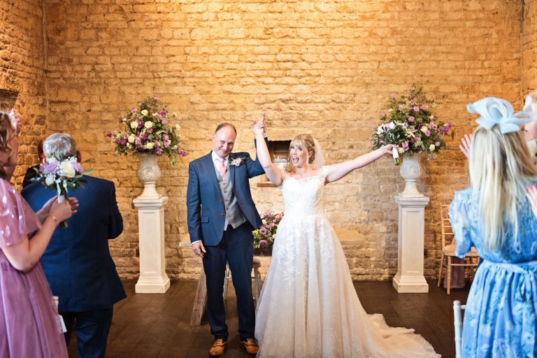 Bride raises her hand in the air whilst holding her new husbands hand. Guests congratulate them.