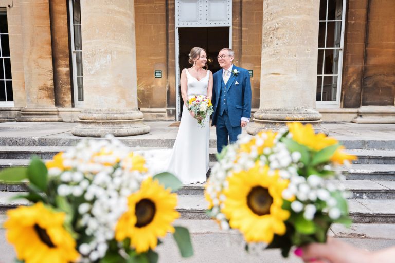Bride and her dad stand on the steps of Leigh Court, the wedding bouquets (with sunflowers) frame the picture.