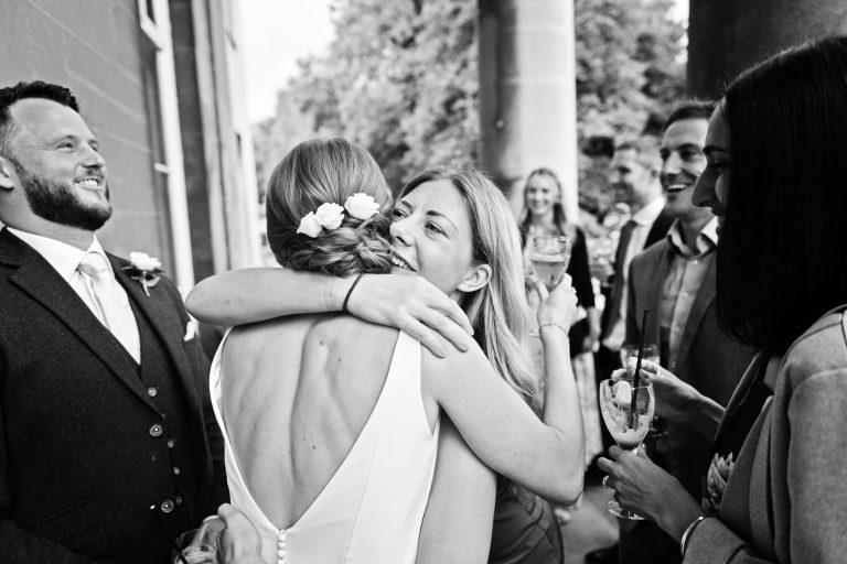B&W photo of a bride being hugged by a friend surrounded by friends and family.