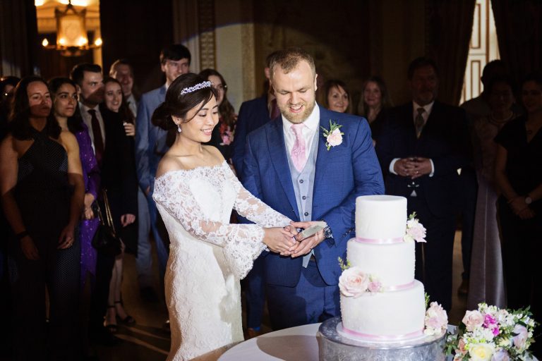Bride and groom cut their wedding cake with friends and family watching behind.