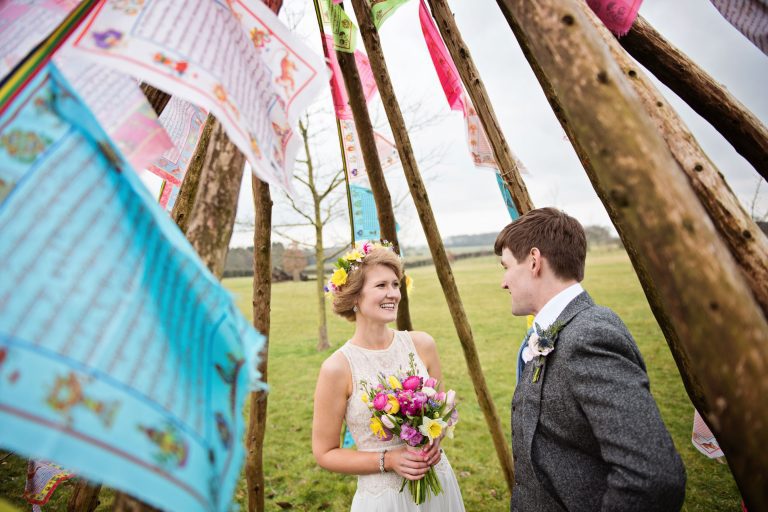 Bride and groom stand in the middle of a teepee structure decorated with asian prayer flags.