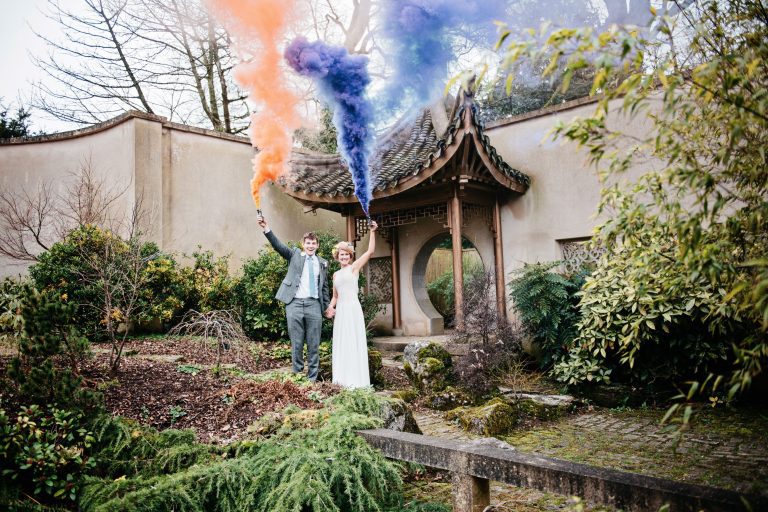 Bride & groom standing, smiling, holding two smoke grenades both lit with orange & purple smoke coming out. The couple are stood in a Japanese Garden in the Matara ceremony gardens. A Japanese wooden archway is behind them.,
