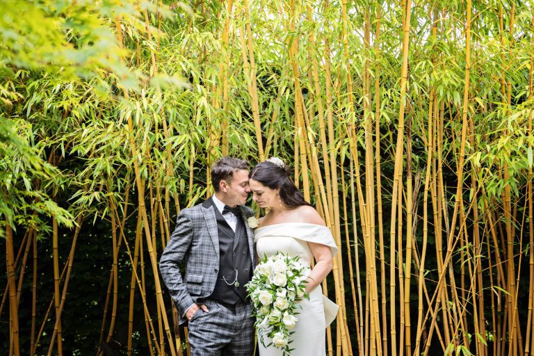 Bride and groom standing, with their heads together, looking content. The couple are standing in front of large bamboo.