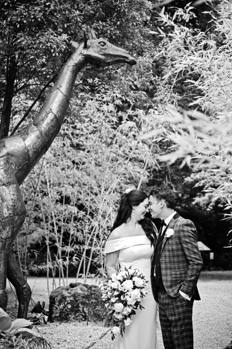 B&W portrait photo of bride & groom standing, with their heads together, smiling. Intimate. However a large sculptured giraffe stands above them.