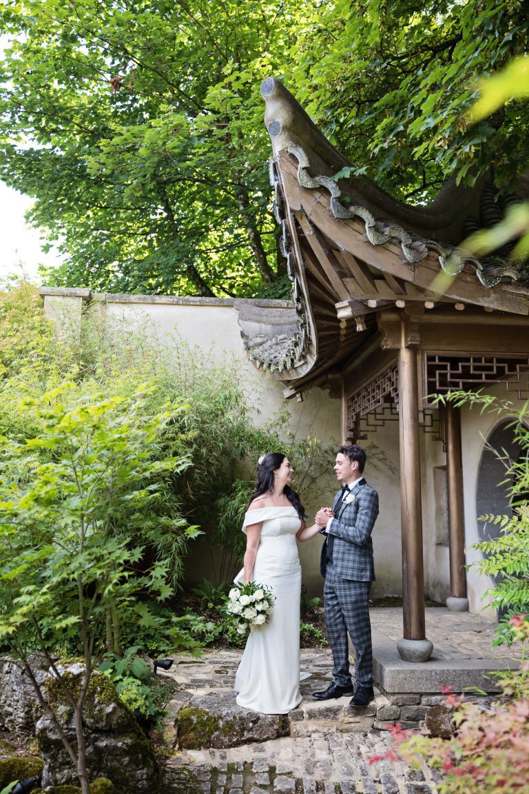 Bride and groom hold hands and talk. Japanese style ornate wooden structure behind them. Also surrounded by acers, bamboos and maple trees.
