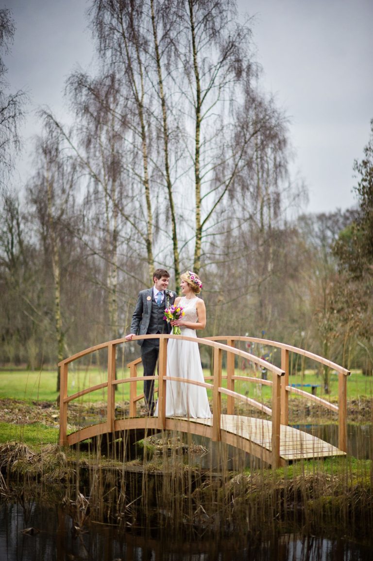 Bride and groom stand on the bridge over the lake and gardens on a late winters afternoon.