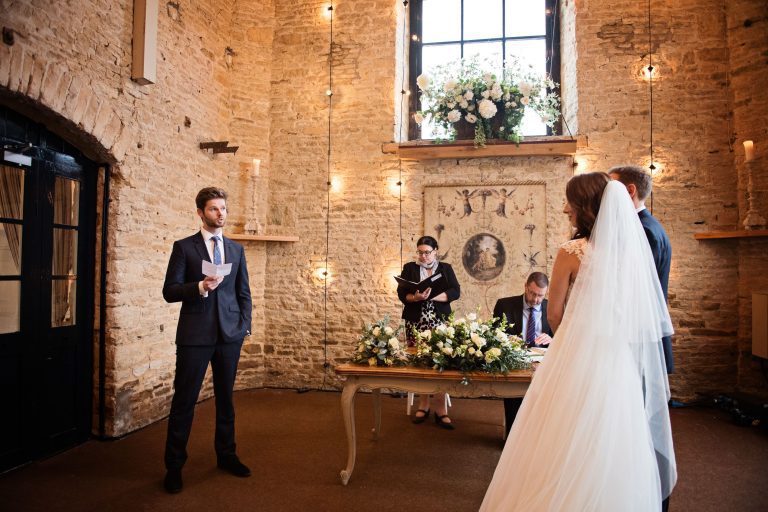 Wedding ceremony at Merriscourt. A friend makes a reading towards the bride and groom. Stone walls and lights and wedding flowers and old art make this look magical.