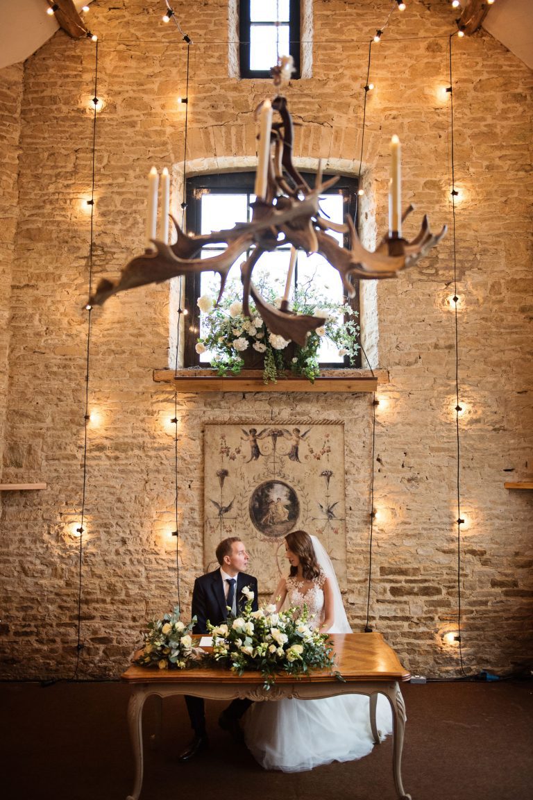 Portrait photo of bride and groom sitting whilst they sign the wedding register. Stone walls and lights and wedding flowers and old art make this look magical.