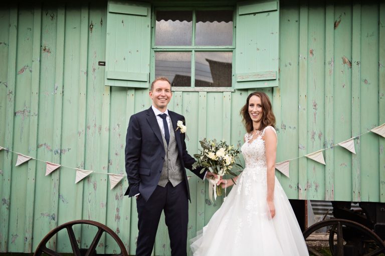 Bride and groom hold the wedding flowers and stand in front (smiling facing the camera) of a wooden pastel green cart at Merriscourt.