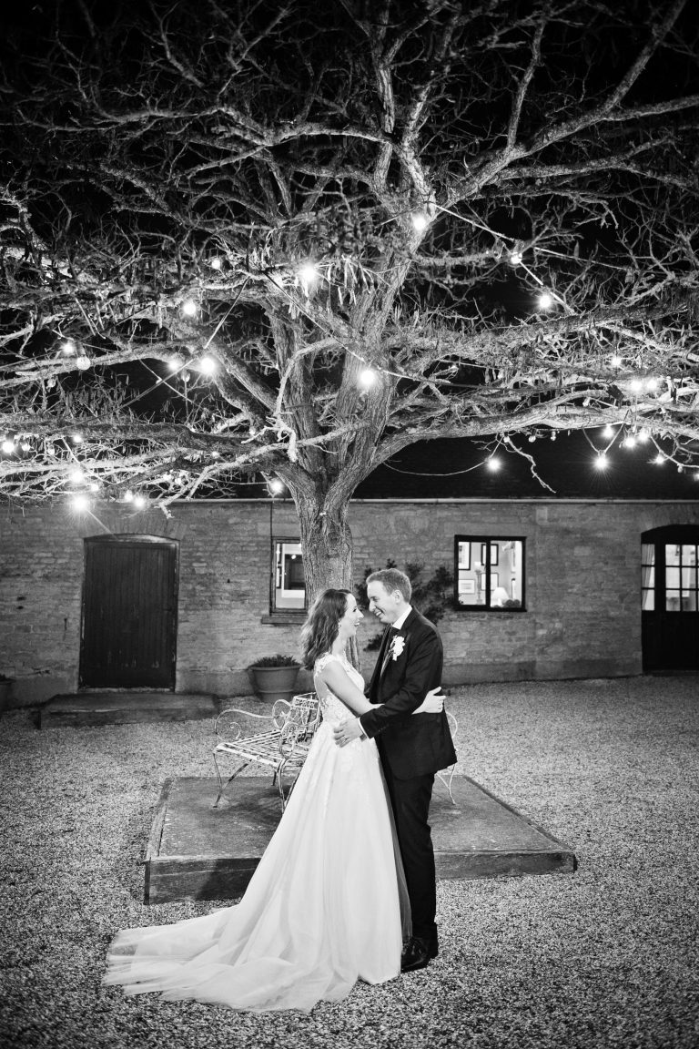B&W portrait photo of bride and groom standing under the famous courtyard fairytale tree at Merriscourt. Photographed at night time so that the tree/fairy lights light up the tree and bride and groom.