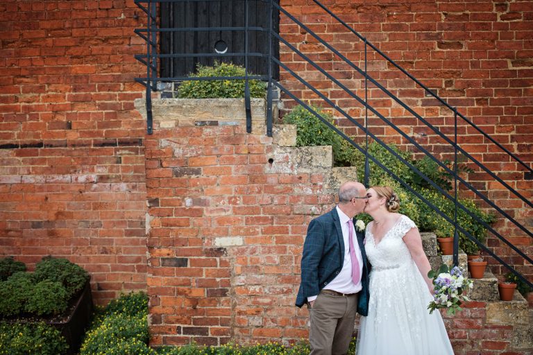Bride and groom kiss with a red brick stairs and wall behind them.