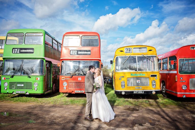 Bride and groom standing front of some colourful old buses in a bus graveyard.