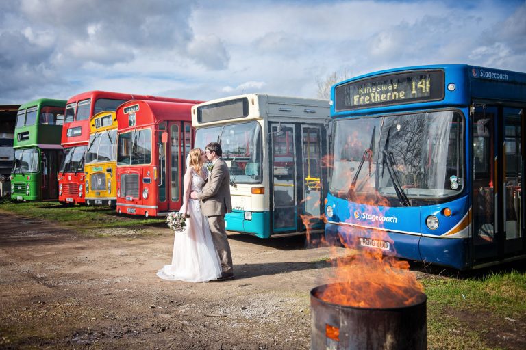 Bride and groom standing front of some colourful old buses in a bus graveyard. A fire is also burning.