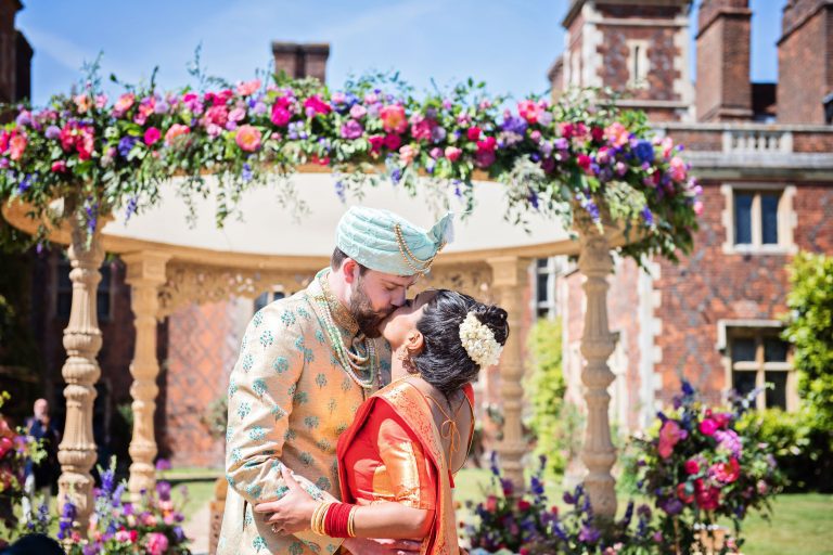 Bride and groom kiss in front of a Hindu ceremony flower arch. Colourful.
