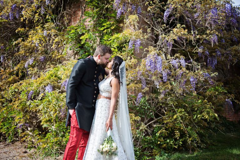 Bride and groom kiss with wisteria climbing plant behind them.