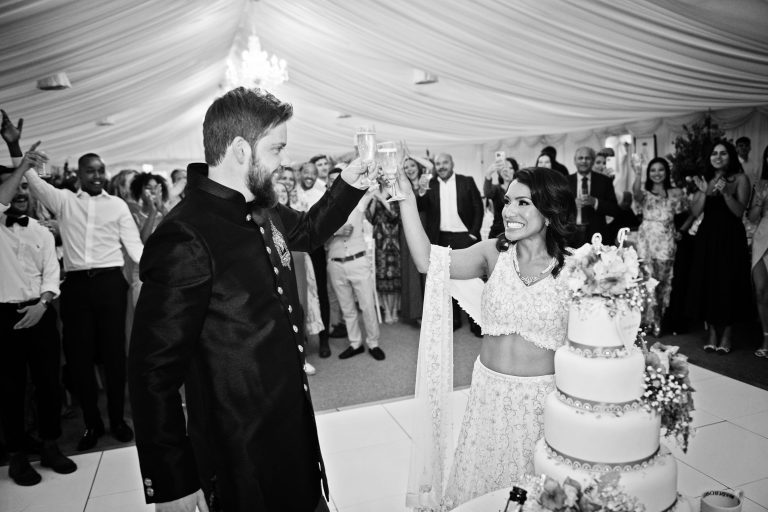 B&W image of bride and groom on the dancefloor, standing in front of their wedding cake and guests. Bride and groom are cheering each other with a glass of fizz.