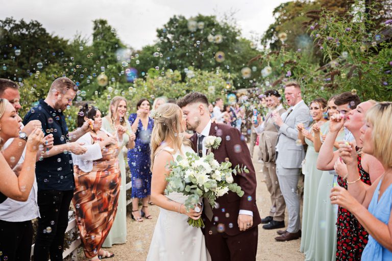 Bride and groom kiss with lots of bubbles and friends surrounding them.