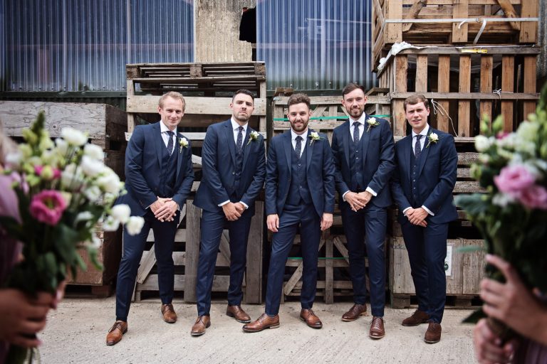 Groomsmen striking a pose with bouquets framing the image. Wooden pallents and a barn behind them.