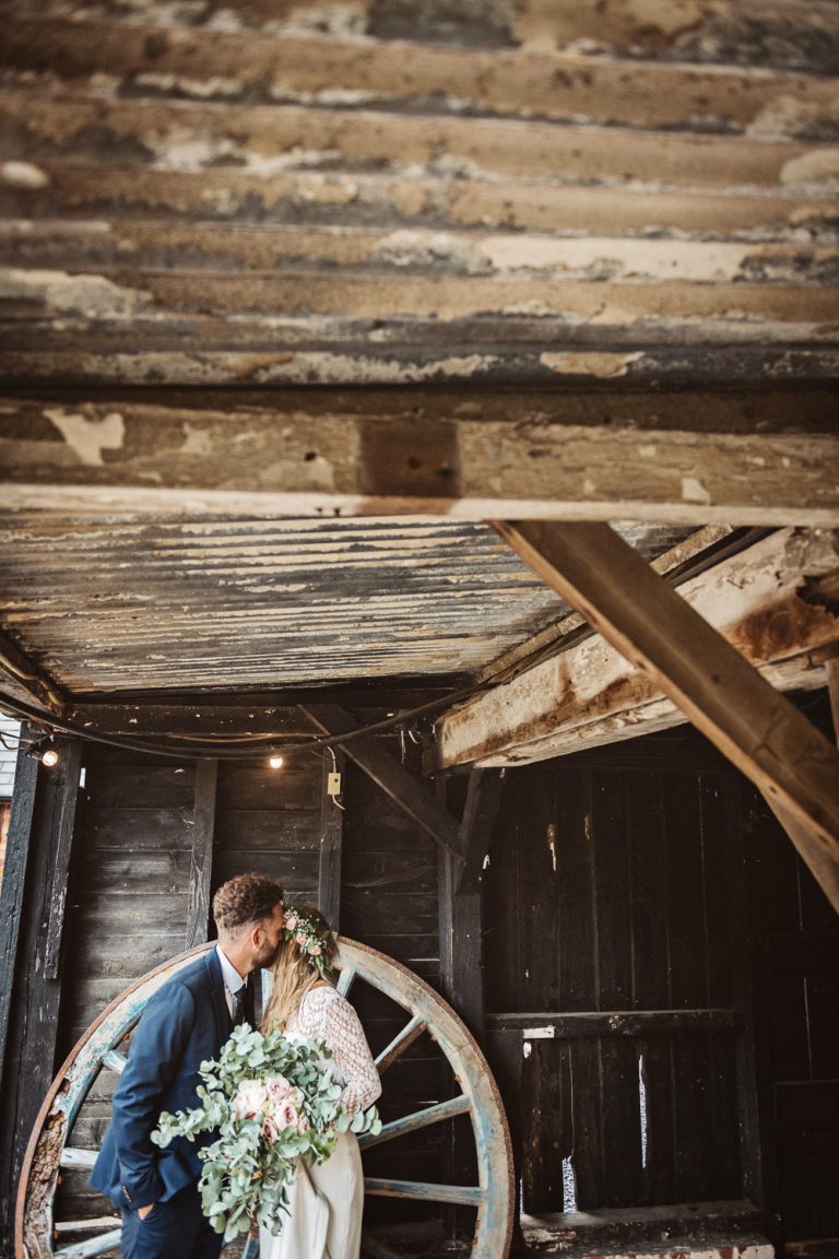 Bride and groom kiss next to an old wooden wheel in a wedding barn.