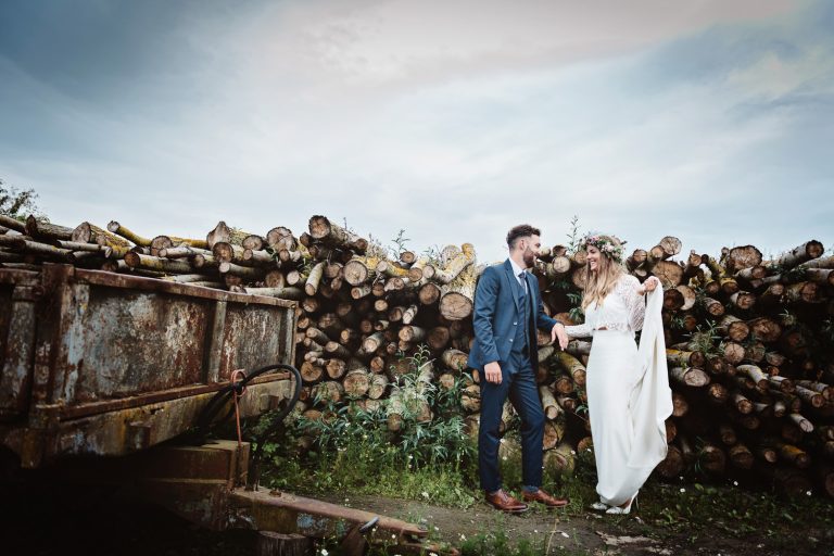 Bride and groom stand next to a rusty trailer and pile of logs.
