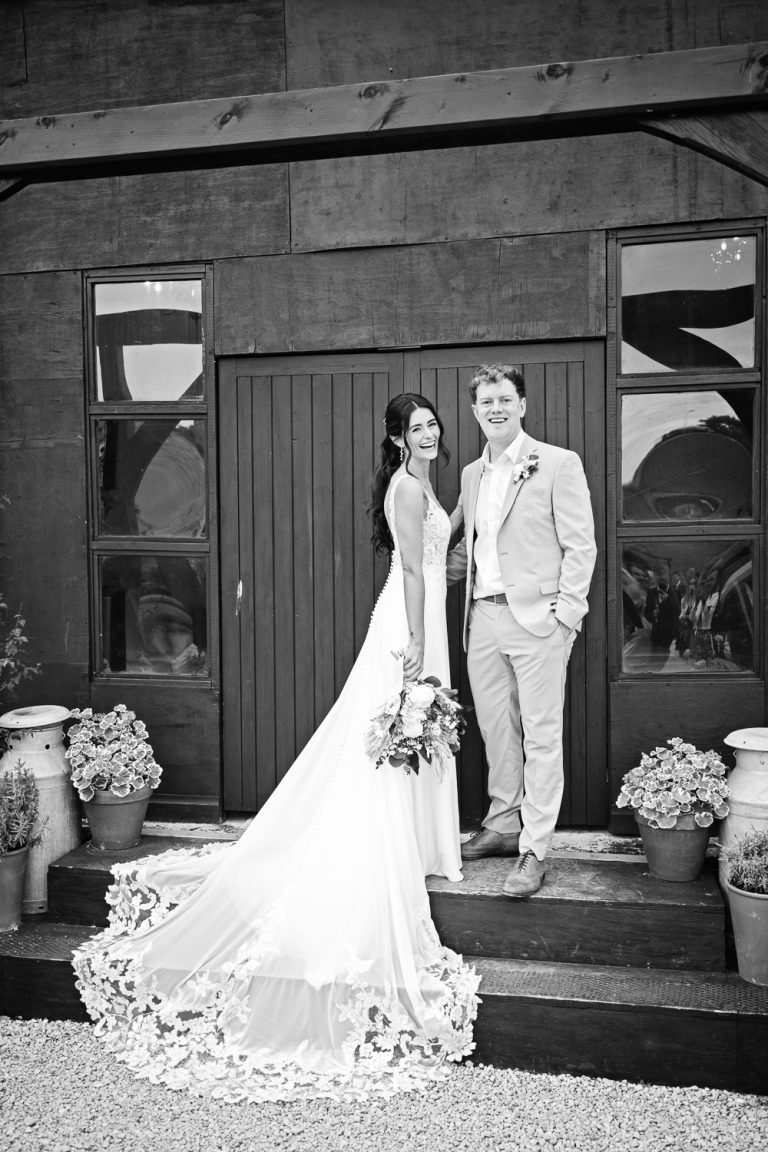 B&W portrait image of bride in her long train wedding dress, standing next to her new husband. Both smiling. Standing on two stairs leading to a wooden threshing barn at Slapton Manor