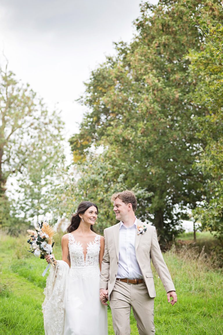 Portrait photo of bride and groom walking together in hand in hand smiling with gorgeous mature trees and green countryside behind them.