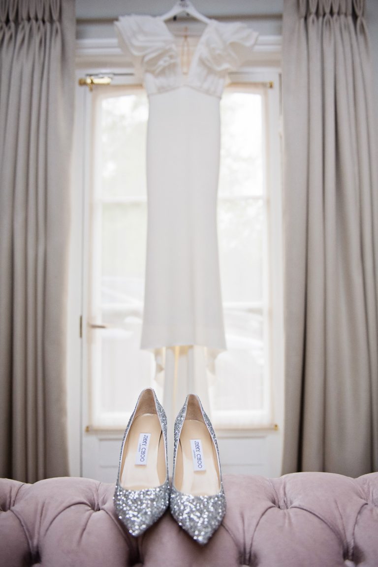 Portrait photo of brides Jimmy Choo wedding shoes perched on a pink/purple sofa. White wedding dress is hung up on the window behind it.