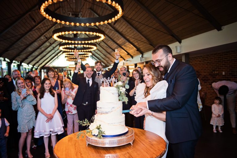 Bride and groom cut their wedding cake at Syrencot with their wedding guests cheering and smiling behind them