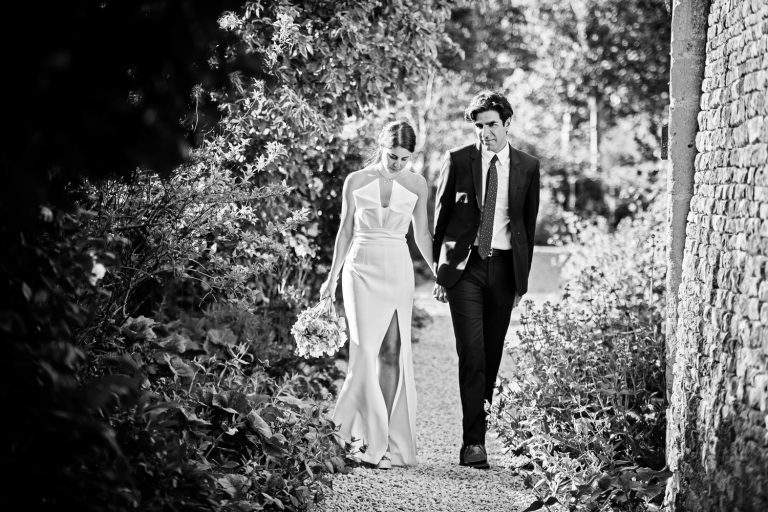B&W candid photo of bride and groom walking hand in hand down a gardne path. Natural.