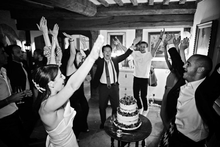 B&W documentary wedding photo of bride and bridal party with their hands in the air celebrating the wedding cake.