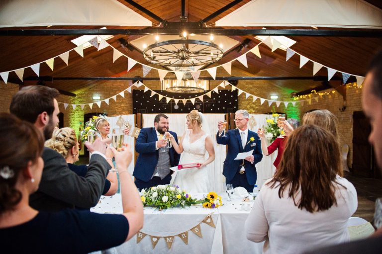 Wedding guests and bride and groom and top table saying cheers to each other whilst raising their champagne glasses, candid photo. Wonderful pastel coloured bunting decorates the scene/barn.