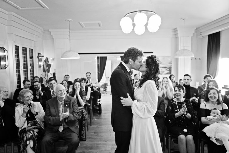 Candid photo of Bride and groom seal their wedding with a kiss. Wedding guests sitting behind them clapping and congratulating them at The Berkeley Square Hotel.