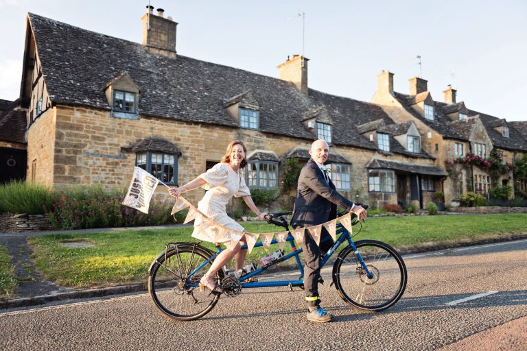 A fun photograph of a bride and groom sitting on their tandem bike in the cotswold village of Broadway during golden hour.