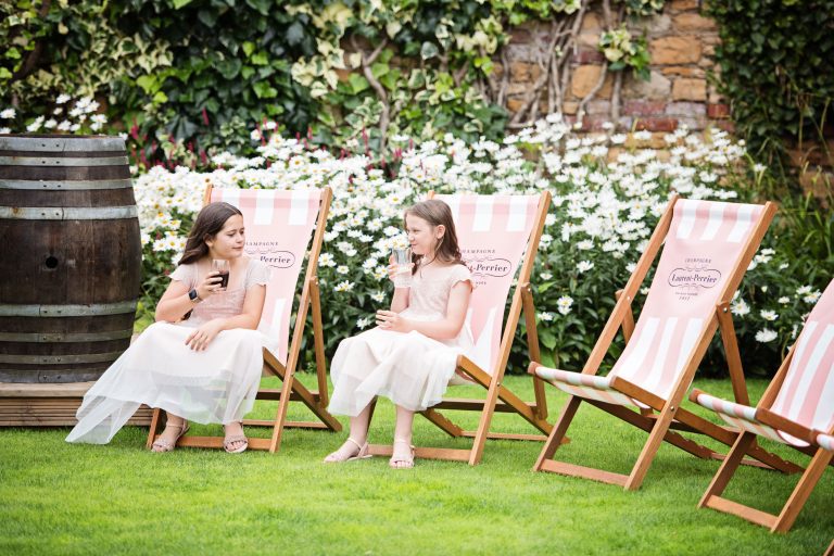 Two flower girls sit on beach style chairs sipping on lemonade and coke in a cotswold garden.