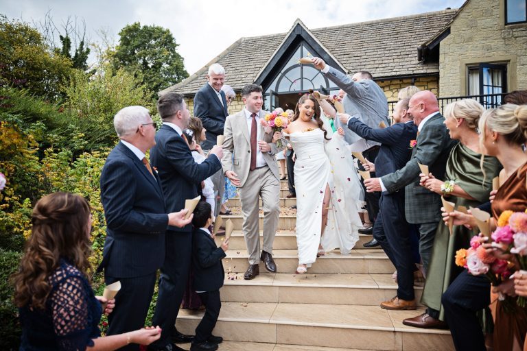 Candid photo of Bride and groom walking down the steps as their wedding guests throw confetti over them.