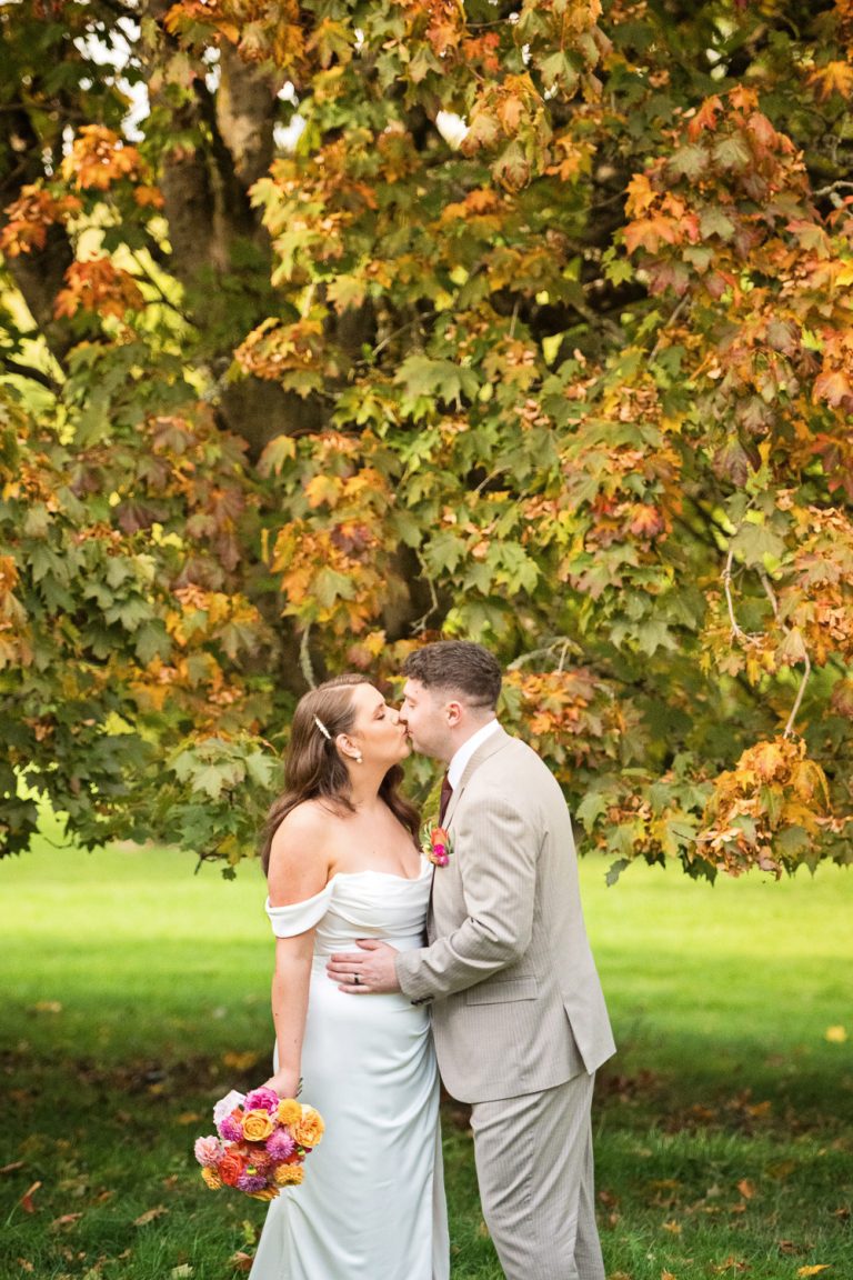 Bride and groom kiss in front of a Autumnal tree that's leaves are turning colour.