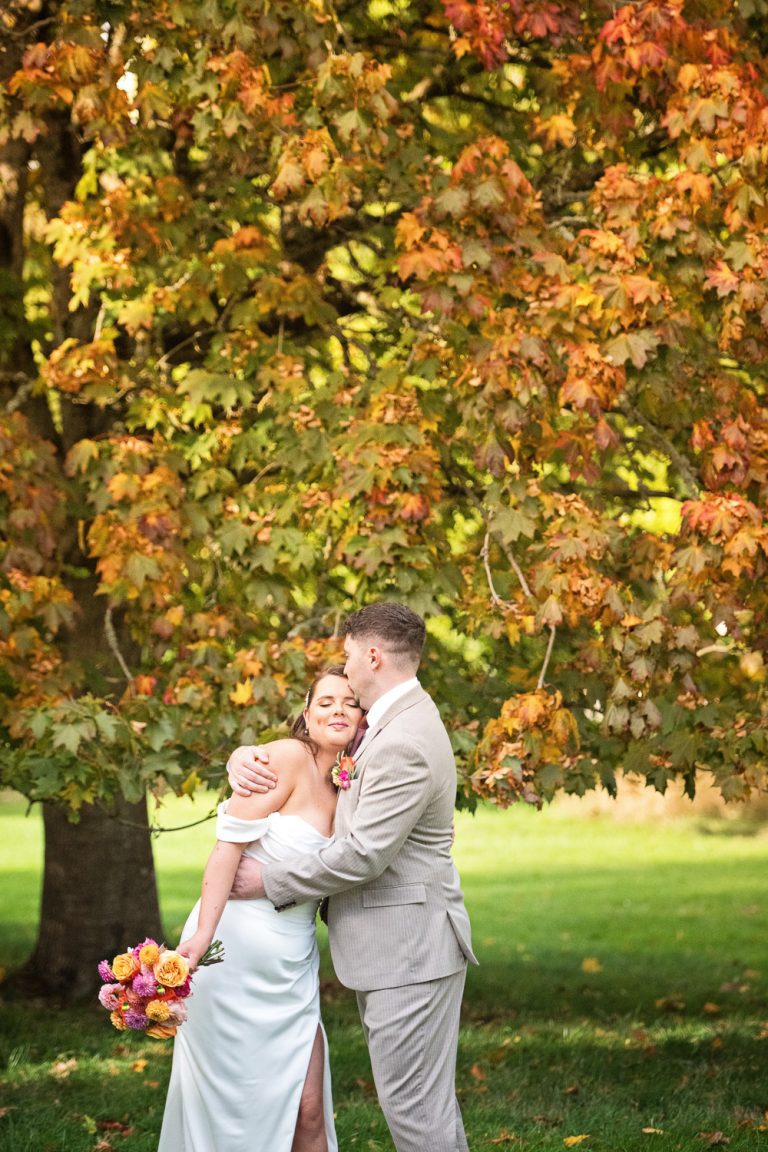 Bride and groom hug in front of a Autumnal tree that's leaves are turning colour.