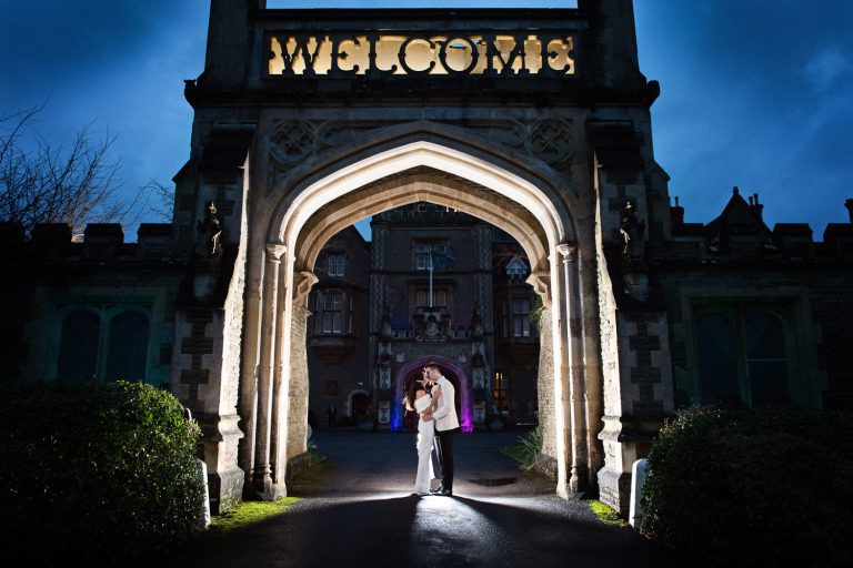 Night time photo of bride and groom kissing under archway entrance at Tortworth Court. Bride and groom are lit up.