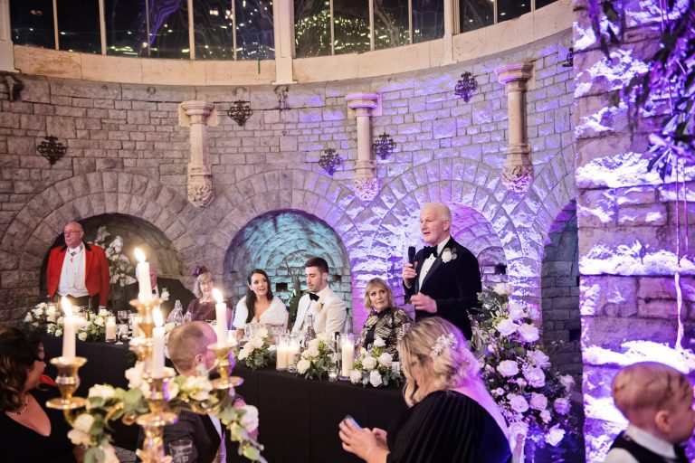 Father of the bride delivering his speech. Bride and top table looking onward at him.