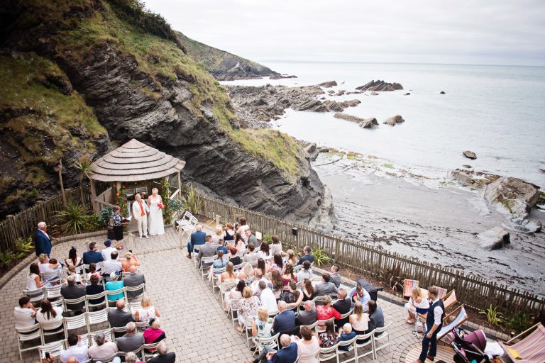 Birds eye view of the wedding ceremony taking place at Tunnel Beach, with guests sitting down and the wedding couple stood at the top of the wedding isle. Impressive view of the Devon coastline.