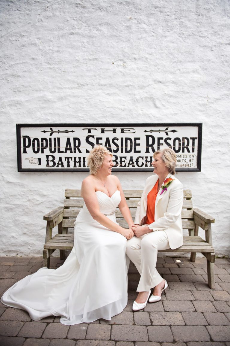 Bride and bride sitting on a bench in front of the sign saying "The Popular Seaside Resort, Bathing Beach."