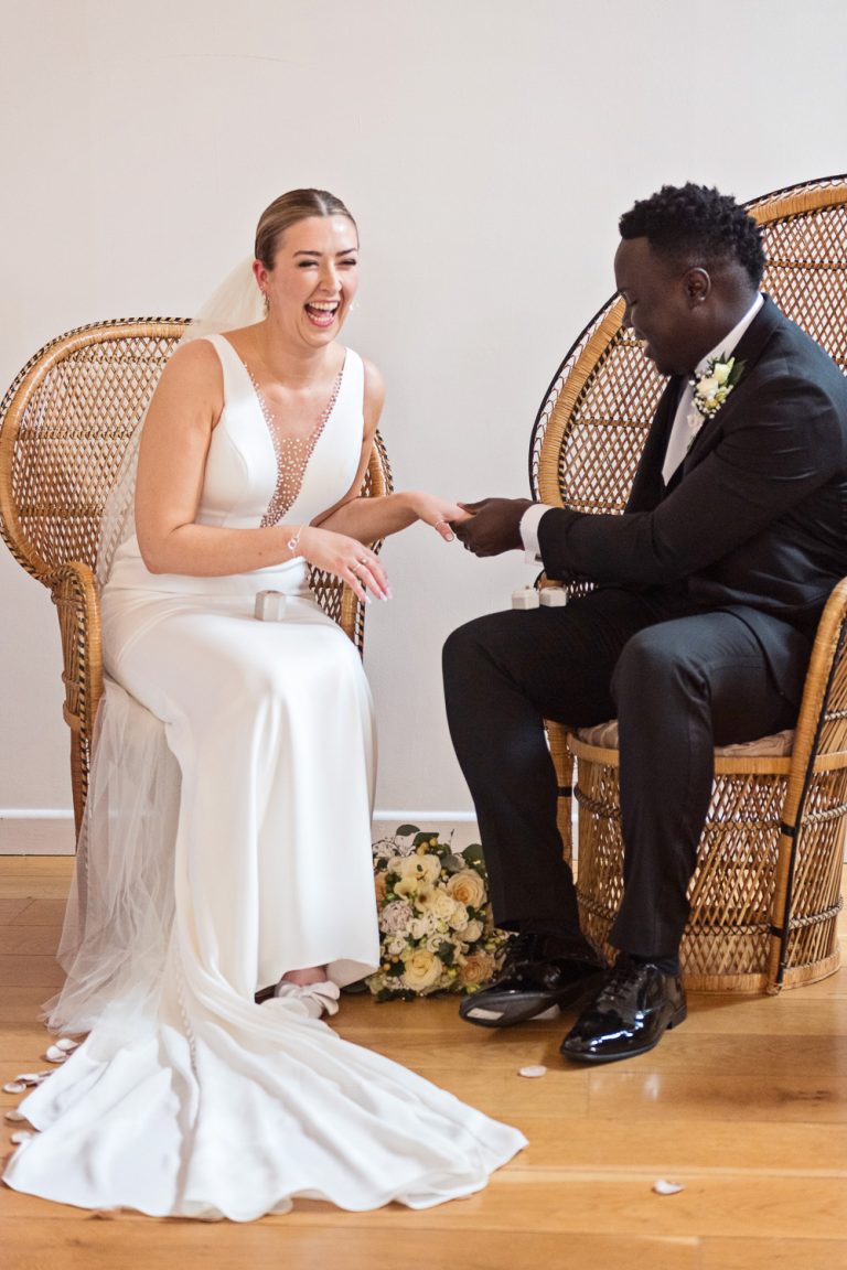 Portrait documentary photo of bride laughing as the groom puts the wedding ring on her finger.