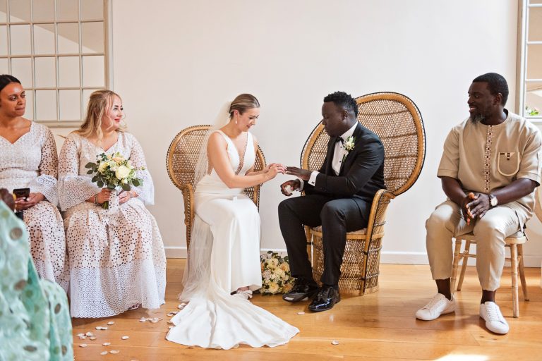 Bride putting the wedding on the grooms finger, watched by close family members.