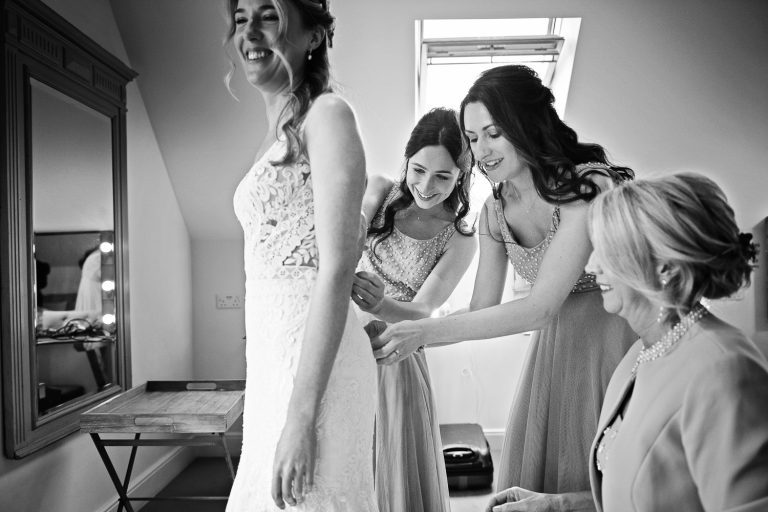 B&W photo of bride in her wedding dress being helped by her bridesmaids.