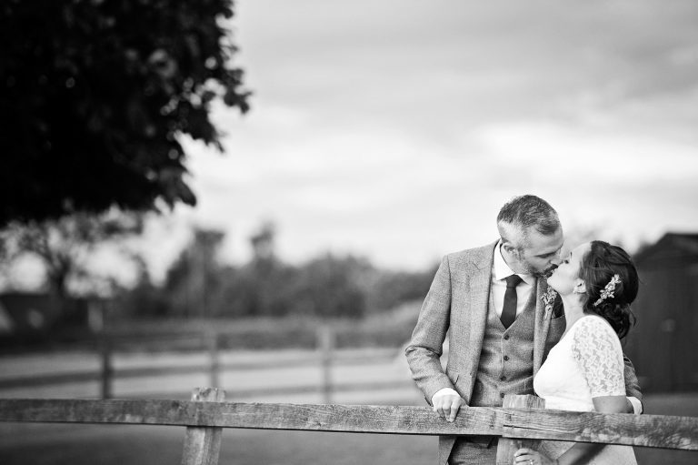 B&W photo of bride and groom kissing up against a post fence.