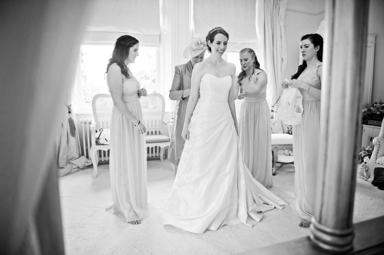 B&W candid photo of the bride getting into her wedding dress helped by the mother of the bride and her bridesmaids.