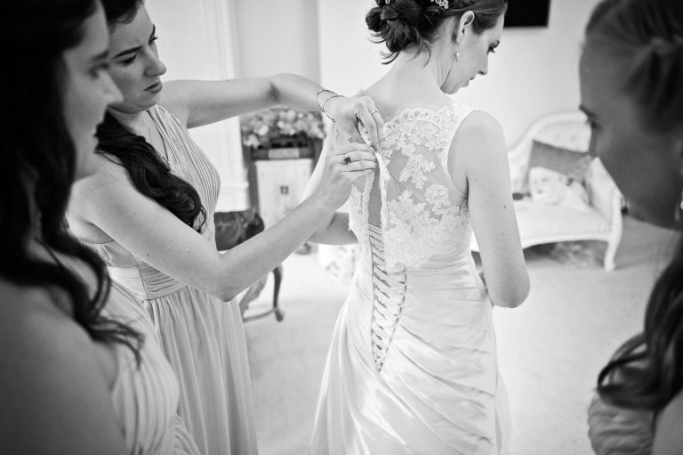B&W candid photo of the bride having the back of her wedding dress done up by her bridesmaids.