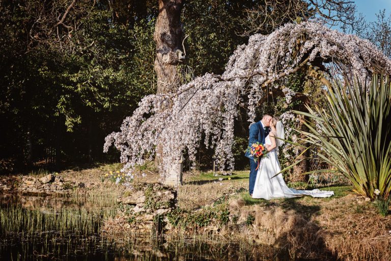 Bride and groom kiss underneath a blossoming tree. They are standing next to a lake.