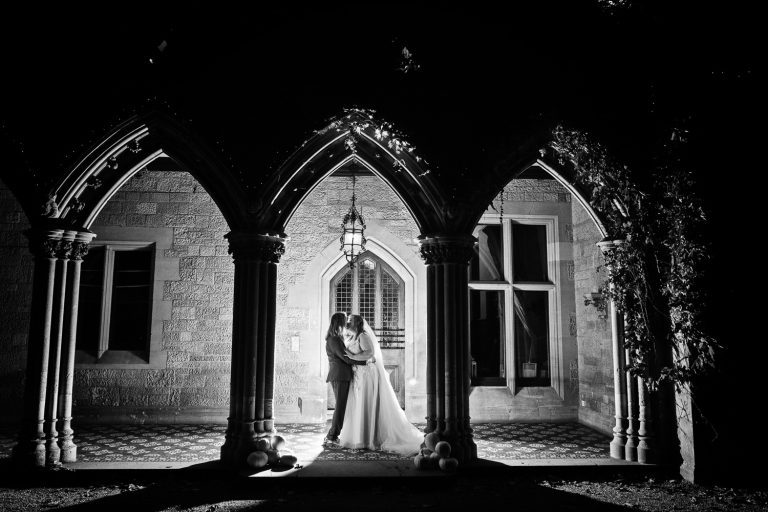 B&W photo of bride and groom kissing in the dark in between the front arches at Manor By The Lake. The pillars are made into silhouettes by the lighting.