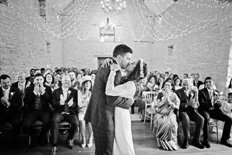 B&W photo of bride and groom kissing after being announced husband and wife, their wedding guests applauding behind them.
